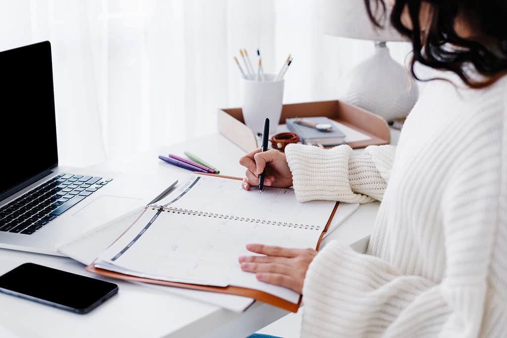 woman at white desk writing in a planner with a white shirt