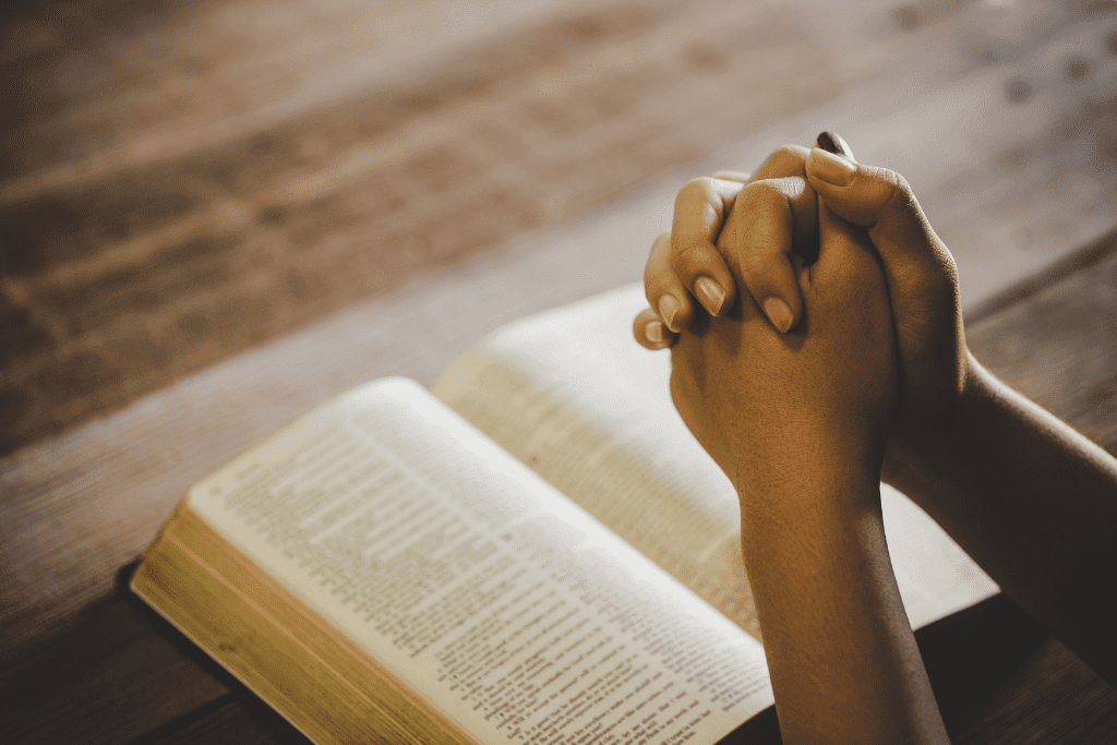 woman praying with folded hands on top of open Bible on wooden desk